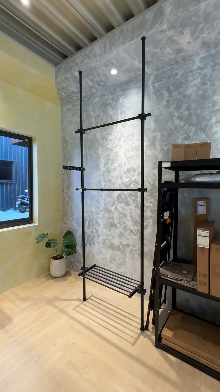 Black steel pipe coat rack with a bottom shelf against a concrete wall.