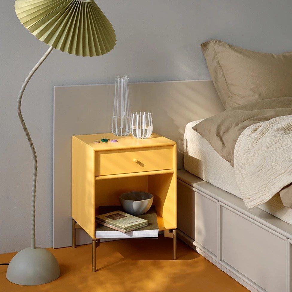 Yellow bedside table with a single drawer next to a beige bed.