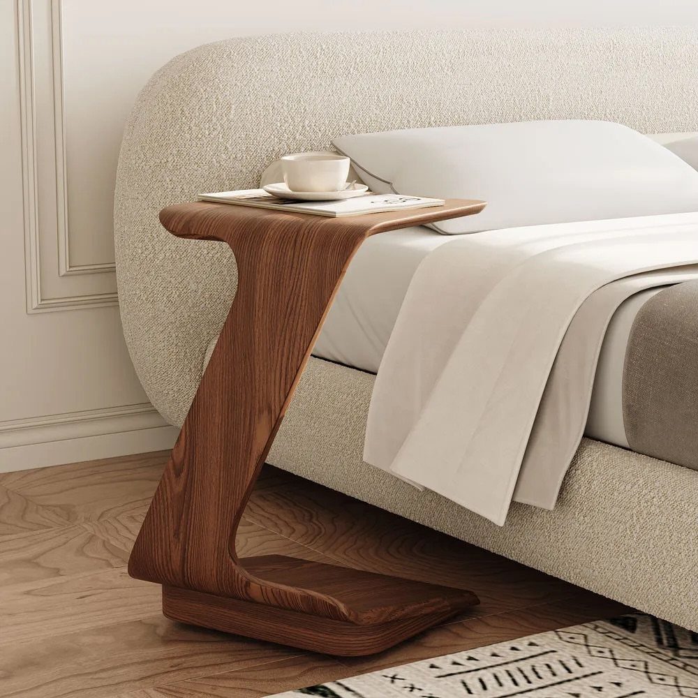 C-shaped wooden bedside table next to a white bed with a white cup.