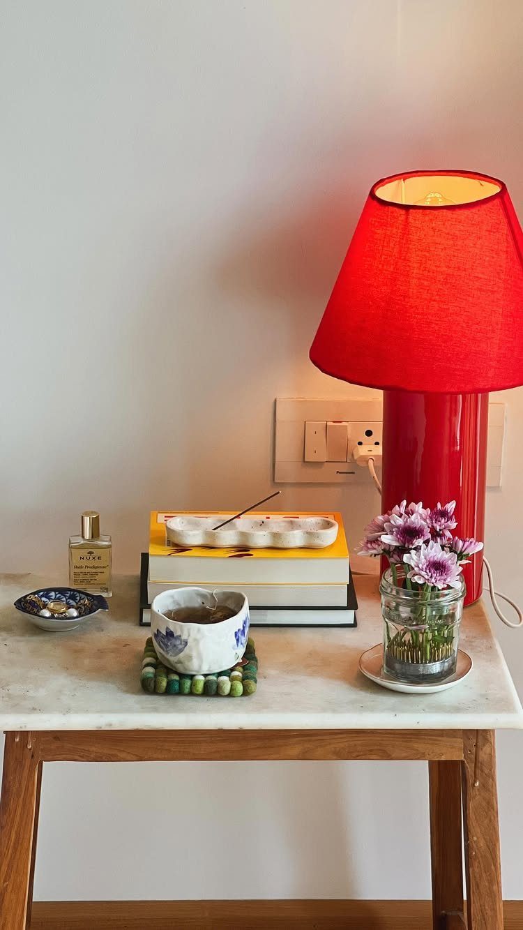 Wooden bedside table with marble top holding a red lamp and purple flowers.