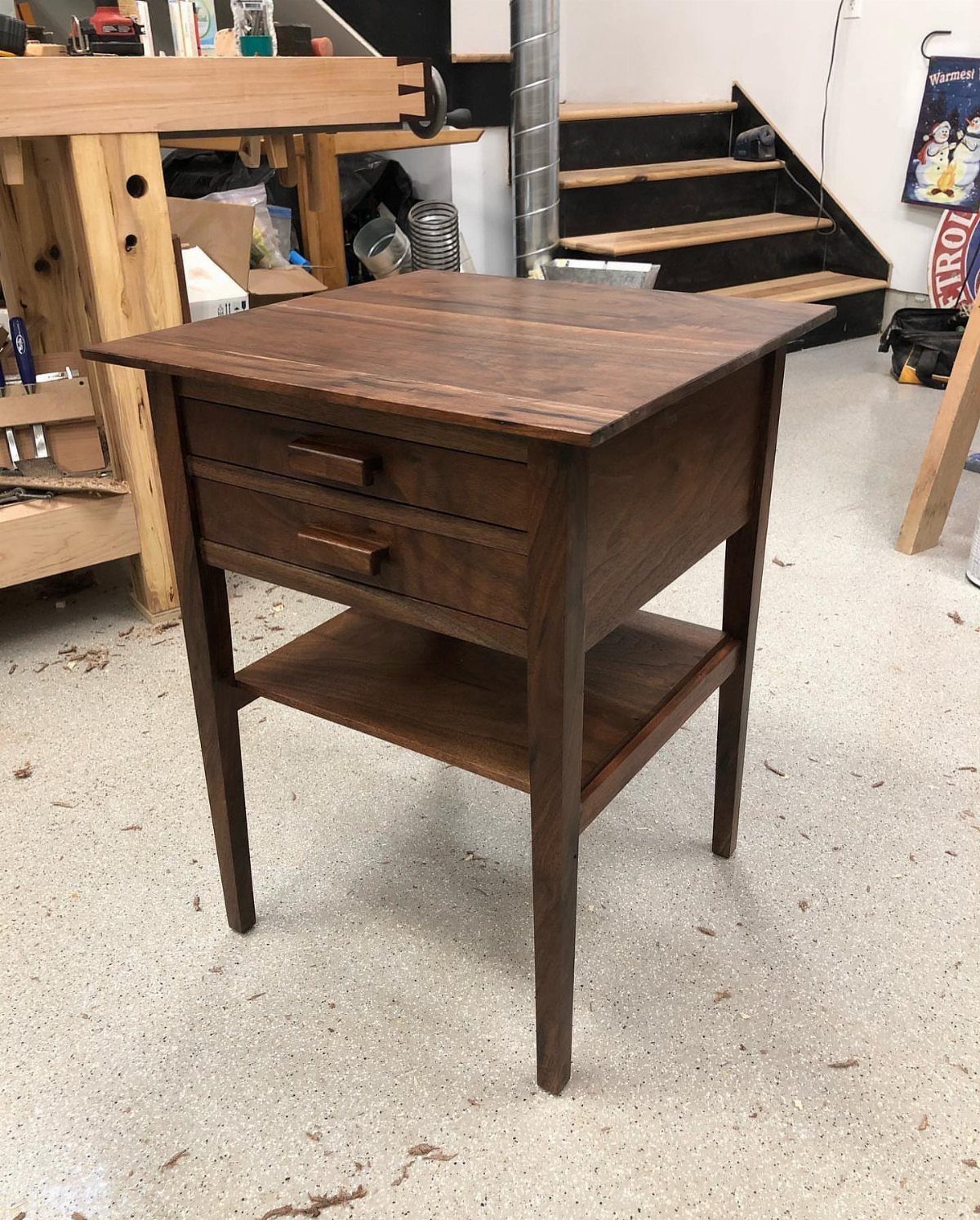Walnut wood end table with two drawers and a lower shelf in a workshop.