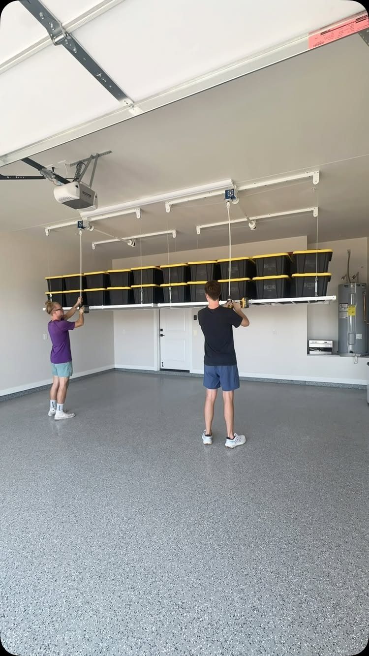 17 Garage Storage Inspiration Ideas to Organize Any Space 14 Ceiling-mounted garage storage system with black and yellow bins being lowered by two people.
