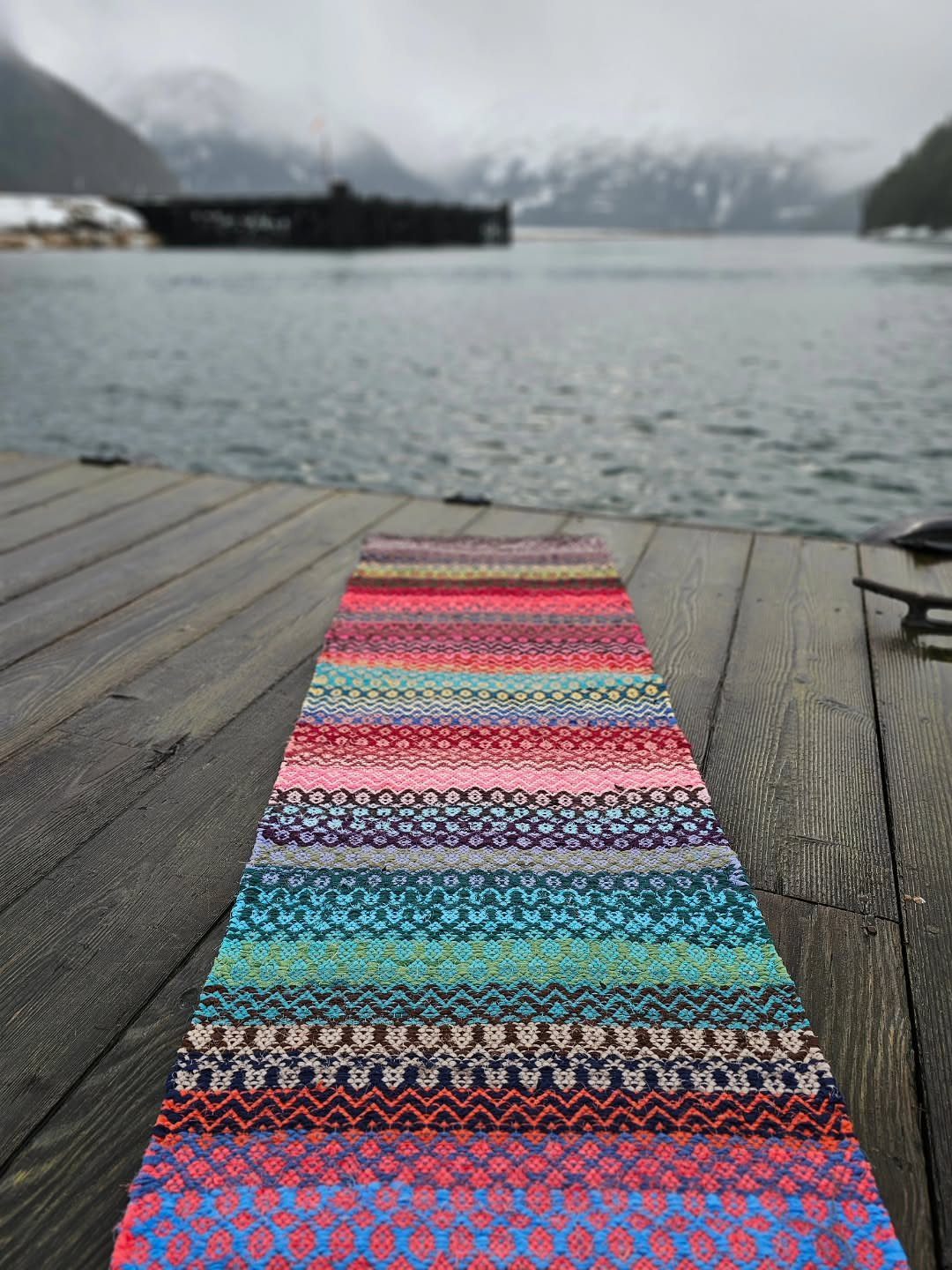 Multi-colored woven rag rug runner on a wooden dock by a misty lake.