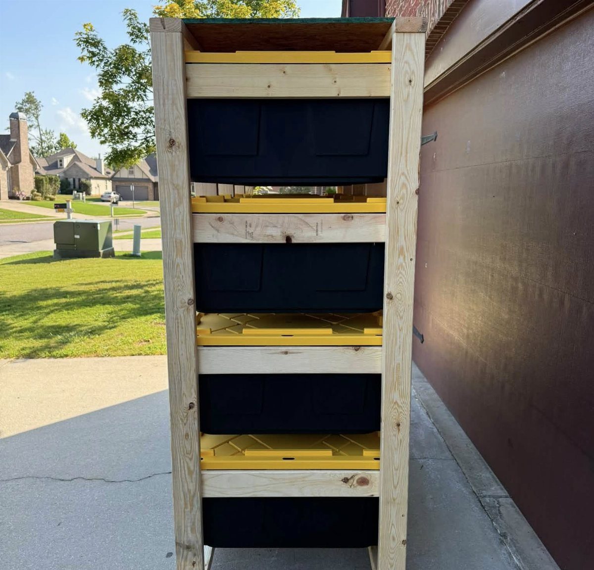 Wooden storage rack holding four black plastic totes with yellow lids outdoors.